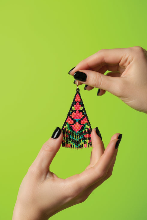 A pair of hands holding a colorful, beaded earring against a bright green background.